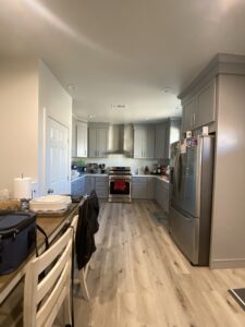 Modern kitchen with gray cabinets and stainless steel appliances, including a refrigerator and a stove. Wooden floor and a table with chairs are visible in the foreground, along with various kitchen items on the countertops.