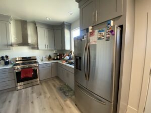 A modern kitchen features gray cabinets, a stainless steel refrigerator adorned with magnets, a gas stove with a red dish towel, and various small appliances on the countertops. Natural light comes in through a window above the sink.