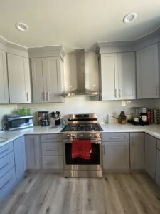 A modern kitchen with gray cabinets, a stainless steel stove and hood, a red towel hanging from the oven handle, and various appliances on the white countertop, including a microwave and coffee maker. The flooring is light wood.