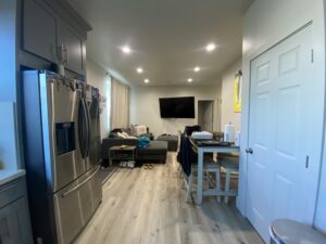 A modern kitchen and living room with gray cabinets and a stainless steel refrigerator. A gray sectional sofa faces a large wall-mounted TV. A small dining table with chairs is on the right, and the floor is light wood.