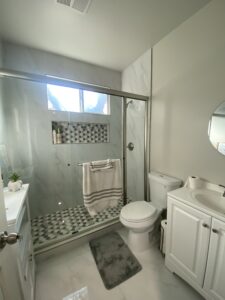 A modern bathroom featuring a glass-enclosed shower with patterned tiles, a white toilet, a white vanity with a round mirror, and light gray walls. A folded towel hangs on the shower door, and a small rug is placed in front of the toilet.