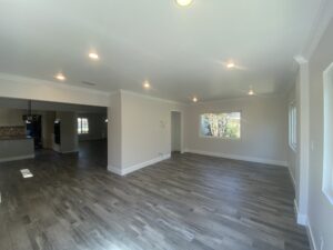 A spacious, unfurnished room crafted by a renowned home builder, featuring gray wooden floors, off-white walls, and recessed lighting. Large windows provide natural light, while an open doorway leads to another room, enhancing the airy and open atmosphere.