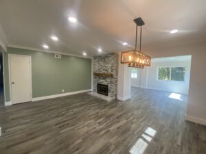 A modern living room crafted by a skilled home builder features wooden flooring, a green accent wall, and a stone fireplace. Recessed lights brighten the space, while a geometric chandelier hangs from the ceiling. Large windows in the background provide natural light.