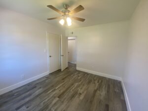 An empty, small room designed by a home builder features light gray walls and wood flooring. A ceiling fan with lights hangs above, with a closed door to the left and an open door on the far side leading to another space. The room is well-lit and simple.