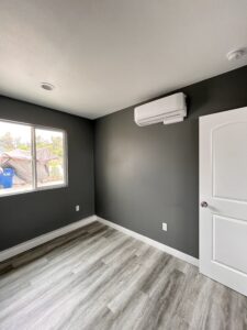 A minimalistic room with gray walls and light wood flooring reflects the craftsmanship of a skilled home builder. A large window on the left lets in natural light, showcasing a glimpse of the backyard. An air conditioning unit is mounted on the wall above a closed white door.