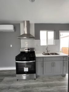 A modern kitchen crafted by expert home builders features a stainless steel stove and hood on the left, adjoining a gray cabinet with a double sink under a small window. The gray walls are complemented by white subway tile backsplash, and light gray wood flooring completes the elegant design.