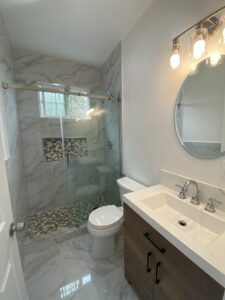 A modern bathroom with a glass-enclosed shower featuring decorative tiles, a white toilet, and a vanity with a sink beneath a round mirror. The space is well-lit with wall-mounted lights, and the flooring is made of marble tiles.