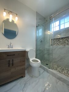 A modern bathroom with a round mirror above a wooden vanity, a white sink, and a toilet. A glass-enclosed shower features marble tiles and a mosaic accent. The floor has light gray tiles, and three light bulbs illuminate the space.