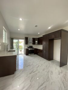 A modern kitchen with dark wood cabinets, a stainless steel oven, and a sleek vent hood. The room features marble-style flooring and countertops, and a sliding glass door leading to an outdoor space, allowing natural light to brighten the space.