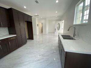 A modern kitchen featuring dark wood cabinets and marble countertops and flooring. The room is well-lit with recessed lighting. A stainless steel sink is installed on the counter. Light is streaming in through a window and a glass door.