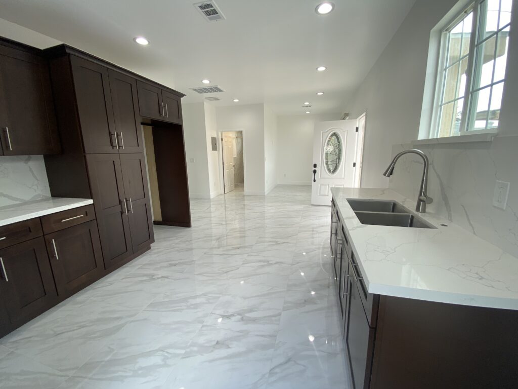 Modern kitchen with dark wood cabinets and a double sink. The room features marble-patterned flooring and countertops. Recessed lighting brightens the space, and a window near the sink allows natural light in. A white door is visible in the background.